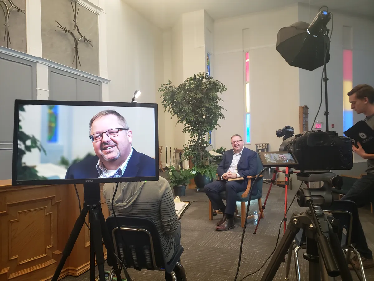 Behind the scenes of an interview in a church sanctuary: the guest seated under a softbox light, his close-up on a monitor in the foreground, and a camera operator at the right.